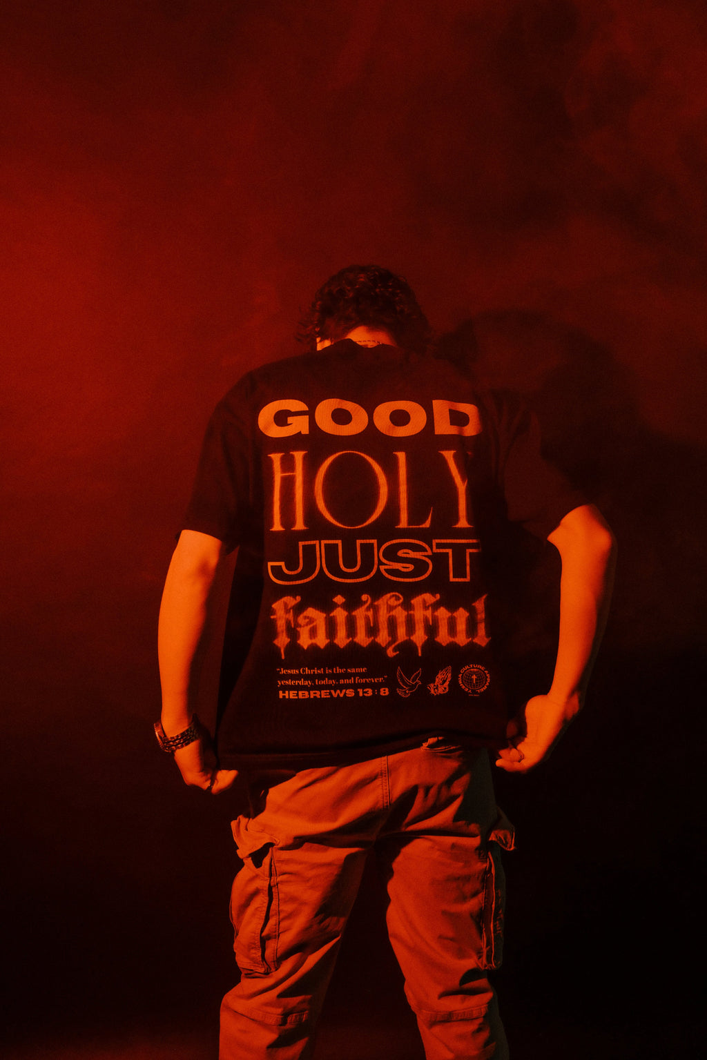Back view of GOD IS tee showing Hebrews 13:8 graphic, red-lit studio