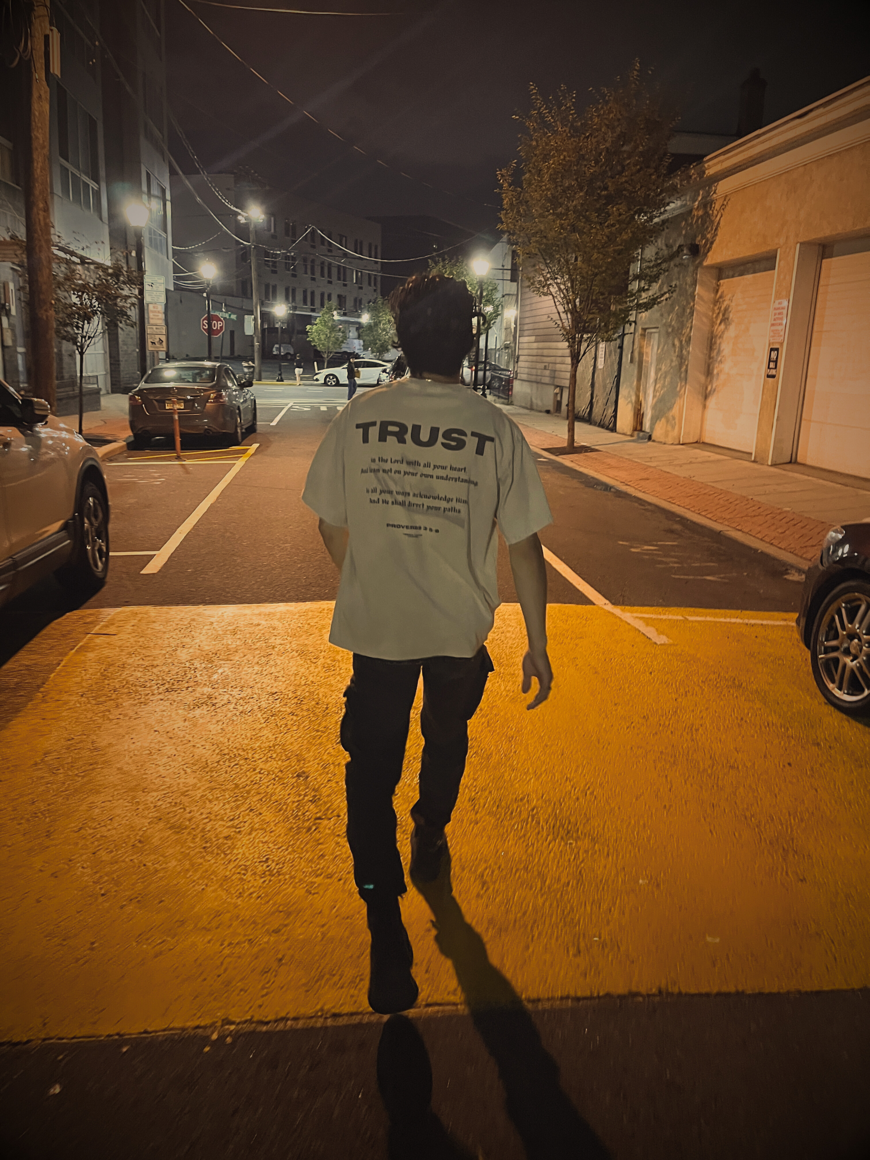 Male model walking at night wearing TRUST Oversized Christian Tee — back scripture Proverbs 3:5–6 visible, urban street scene