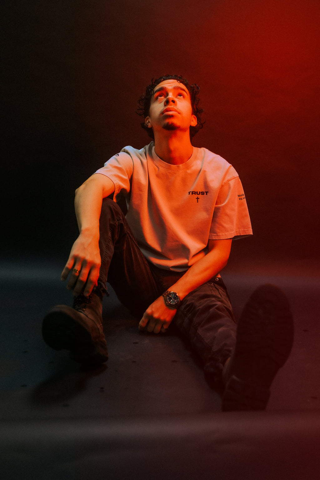 Man wearing TRUST Christian tee, seated pose, red-lit studio shot